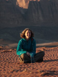 Dr. Jessica Tingle sitting crosslegged in the desert sand with land formation in the background.