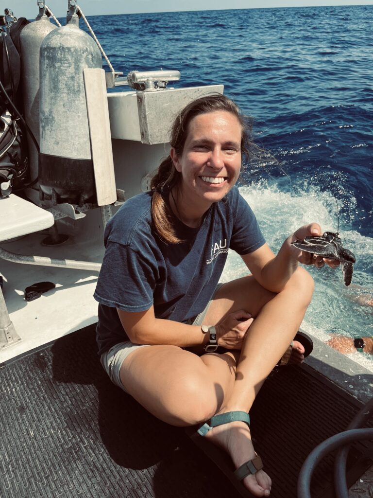Photo of Samantha Trail sitting on a boat holding a turtle with the ocean in the background.